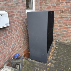 Ein moderner, grauer Schrank steht an einer Backsteinwand, umgeben von herbstlichen Blättern und einem kleinen Tisch.