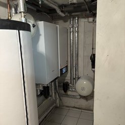 Ein Heizungsraum mit einer modernen Heizungsanlage, Rohren und einem großen weißen Wassertank in der Ecke.