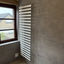 Der schlanke, vertikale Heizkörper aus weißem Metall kontrastiert mit der grauen Wand und dem hellen Fenster.