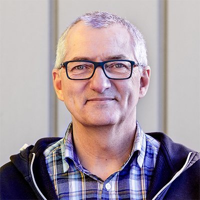 Ein älterer Mann mit Brille und grauen Haaren lächelt freundlich in die Kamera, während er einen karierten Hemd trägt.