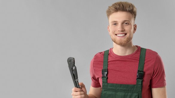 Der Mann in der grünen Latzhose strahlt Freude aus, während er eine Rohrzange präsentiert, vor einem grauen Hintergrund.