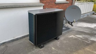 Ein schwarzes Klimagerät steht auf einem grauen Dach, neben einer großen Satellitenschüssel und einer weißen Wand.