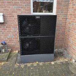 Ein schwarzes Klimagerät mit zwei großen Ventilatoren steht an einer Backsteinwand, umgeben von herbstlichen Blättern.