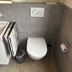 Die schlichte Toilette in einem stilvollen Badezimmer, umgeben von grauen Fliesen und einem Abfalleimer aus Metall.